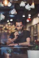 Through window view of positive thoughtful young male enjoying cup of hot coffee while resting in cozy cafe. Pensive bearded hipster guy enjoying morning coffee for breakfast looking through window