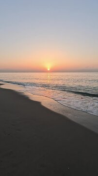 Vertical handheld video of the sun at sunrise or sunset over the horizon of the calm Mediterranean Sea, with clear sky at golden hour, seen from the beach with the waves coming over the sand

