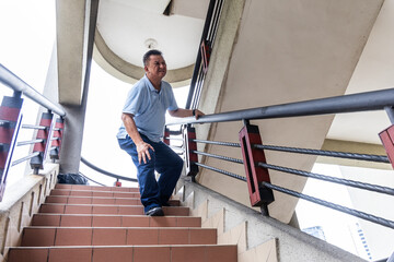Fototapeta premium Elderly Asian man with extreme knee pain holding onto railing carefully attempting to go down staircase