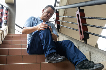 Asian man with extreme knee pain resting on staircase with hands on knees to relieve pain.