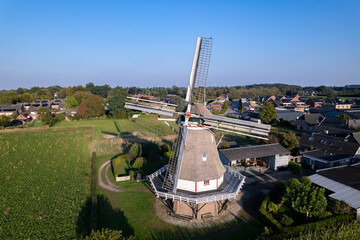 Fototapeta premium Aerial view of traditional windmill in Dutch agriculture fields and flat landscape with village and forest in the background against a blue sky with clouds