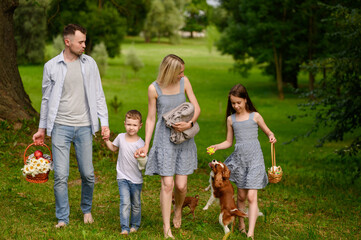 Parents with son and daughter walk together barefoot to park with their pets, blanket and basket with snacks. Cheerful family with children go to picnic in park. Family weekend together.