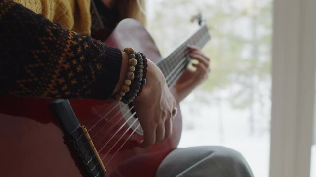 Side midsection shot of anonymous musician with wooden bead bracelets on wrist and in warm christmas sweater playing red acoustic guitar near panoramic window indoors in countryside