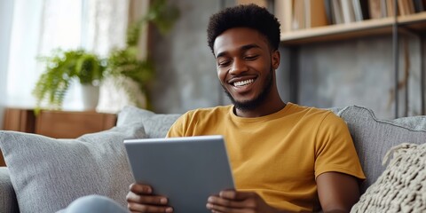 Relaxed smiling african american man holding digital tablet computer using apps sitting on couch at home. Black guy remote learning, social distance working, ordering buying online or reading e book.