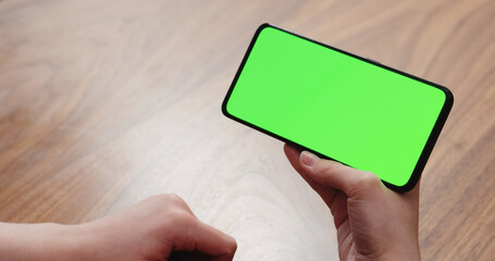 Young woman sitting at a table and using a smartphone with horizontal green screen