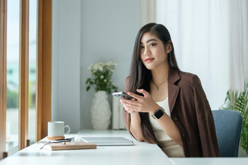 Woman with long dark hair is seated at a sleek, modern desk, looking confidently at the camera while holding a smartphone.