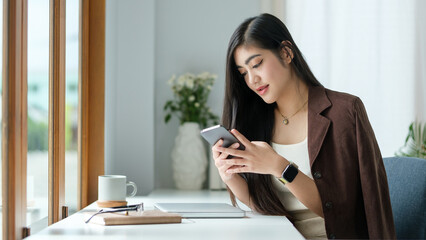 Woman with long dark hair is sitting at a bright, modern desk by a large window, holding a smartphone and smiling as she engages with her device.