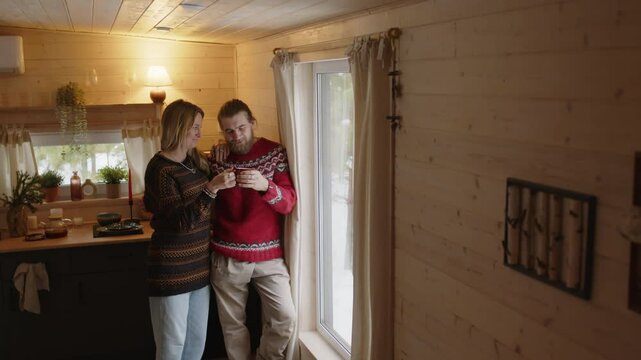 Full medium shot of middle-aged Caucasian couple in warm clothes happily looking out window at winter scenery, wife bringing cup of hot tea to husband, spending time in cottage on christmas vacation