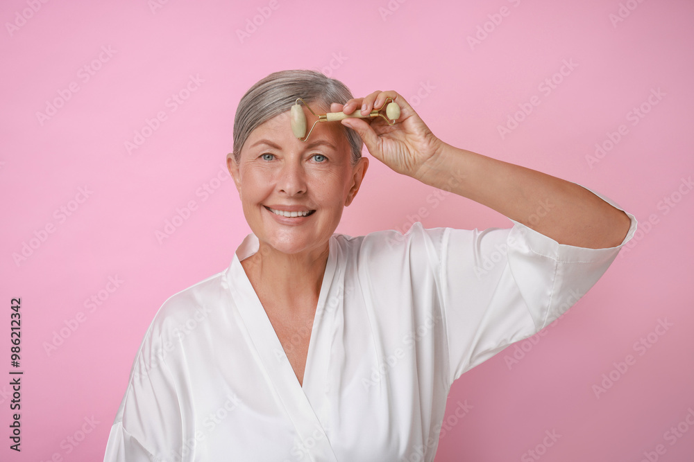 Wall mural Beautiful woman doing facial massage with roller on pink background