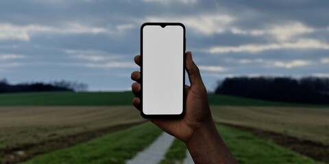 Black African-American male using smartphone with a blank white screen. Gravel road, agricultural fields, cloudy sky apps mockup. Vertical phone orientation