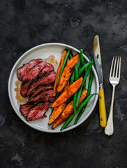 Medium-rare beef steak, sweet potato, string beans - a delicious lunch on a dark background, top view