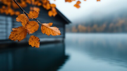 Obraz premium A close-up shot of orange autumn leaves on a branch with a lake and a wooden house in the background shrouded in mist, capturing a tranquil autumn scene.