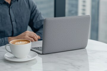 Person working on laptop with coffee cup on marble table, modern office view in background.
