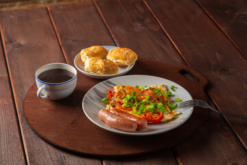 Eggs with tomatoes and sausages in a plate on a wooden table of coffee and croissants on a round stand.