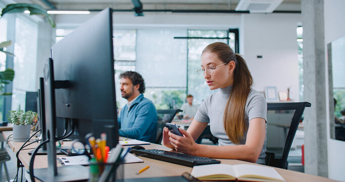 Beautiful girl spending time on phone while sitting at workplace in office. Corresponding with clients or checking her mail. Using computer and smartphone for work efficiency. Modern office.