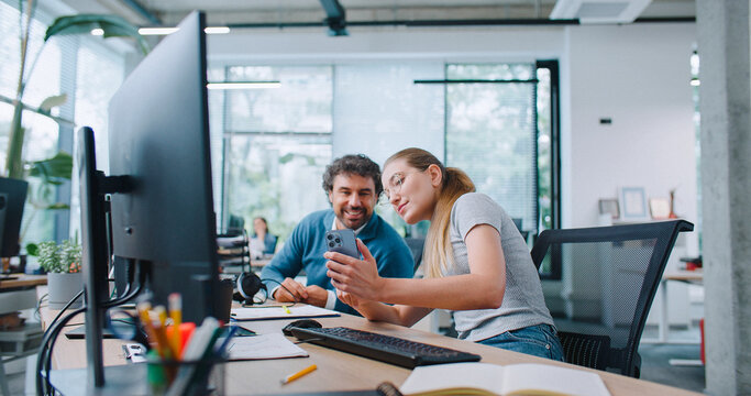 Employees distracted from work by phone. Woman in glasses browsing social networks and showing video or photo on her cell phone to colleague. People cheerfully smiling while sitting at desks.