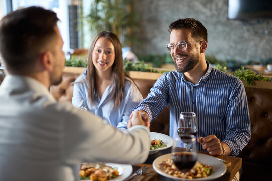 Happy businessmen shaking hands on a casual meeting at a restaurant while their female colleague is next to them.