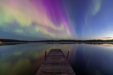 A beautiful Northern Lights is shown in the night sky, with vibrant colors such as pink, green, and purple.  In the foreground, there is a wooden pier jutting out into the water.