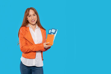Young Woman in Orange Jacket Smiles With Boarding Pass and Travel Guide Against a Blue Backdrop
