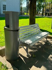 A park bench next to a trash can in a sunny outdoor setting