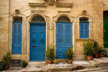Fototapeta premium Charming facade with vibrant blue wooden doors and shutters, set against traditional stone walls in Malta. Potted plants add a touch of greenery to this picturesque Mediterranean scene