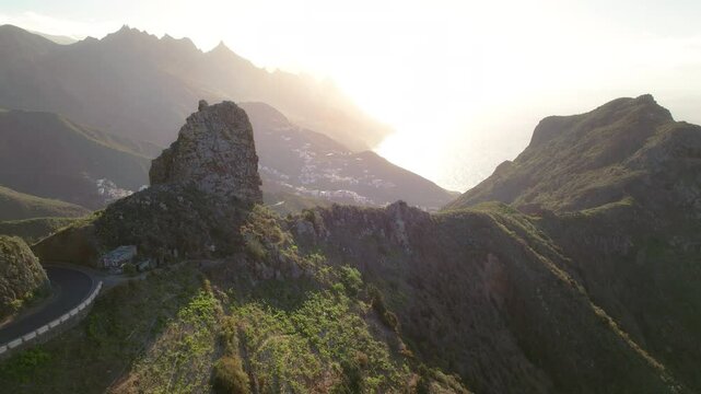 An impressive rocky cliff framed by the golden sunlight, overlooking a small town by the sea. Roque de las Animas in Anaga rural park Tenerife
