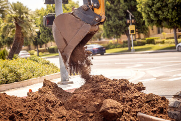 Close up of a backhoe scoop bucket filled with dirt  that is pouring out ontop a dirt pile. The...