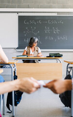 Vertical shot of young teenage boy giving cheat sheet to classmate during examination in classroom....