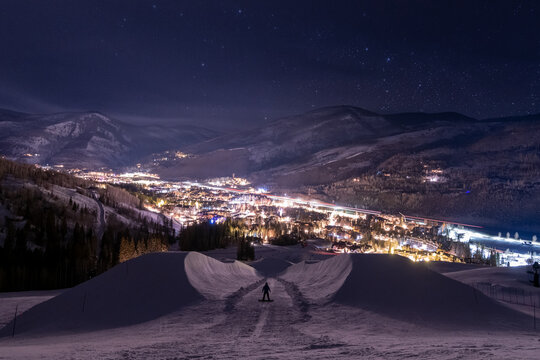 A snowboarder standing on a snowy half pipe on a mountain overlooking the town below