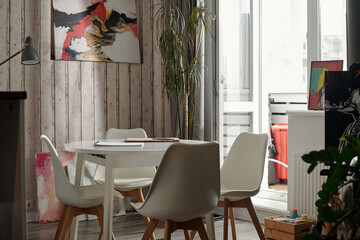 Empty dining table with two chairs placed beside small balcony inviting for peaceful ambiance. Room decorated with wooden wall and abstract paintings creating artistic vibe