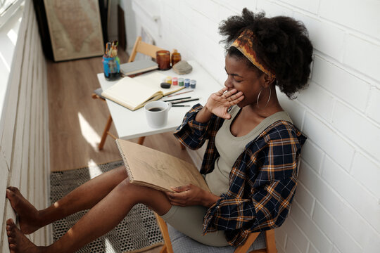Young woman sketching in cozy, well-lit room with artistic supplies surrounding her, enjoying relaxed atmosphere while engaging in creative process