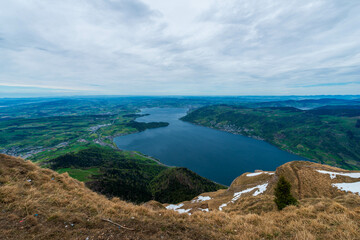 A Swiss Alps vista reveals a stunning panoramic view of Lake Lucerne nestled amidst rolling green hills, framed by a dramatic sky with swirling clouds.