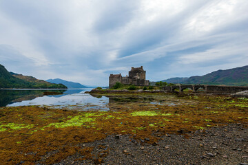 Eilean Donan Castle, a majestic Scottish castle, sits on a small island, its reflection mirrored in the calm waters of Loch Duich, surrounded by a rugged shoreline and dramatic cloudy skies.