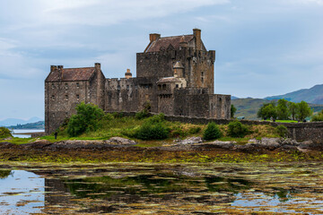 Eilean Donan Castle, a historic Scottish castle, stands majestically on a small island at the confluence of three lochs, surrounded by serene waters and a backdrop of rolling hills under a cloudy sky.