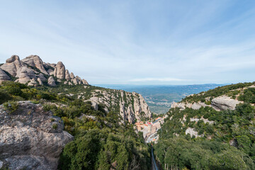 A green funicular railway ascends a steep, forested valley in Montserrat, Spain, with dramatic rocky peaks and a distant view of the valley floor below.