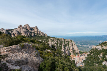 Naklejka premium A green funicular railway ascends a steep, forested valley in Montserrat, Spain, with dramatic rocky peaks and a distant view of the valley floor below.