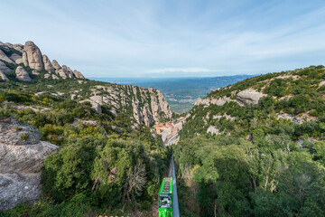 A green funicular railway ascends a steep, forested valley in Montserrat, Spain, with dramatic rocky peaks and a distant view of the valley floor below.