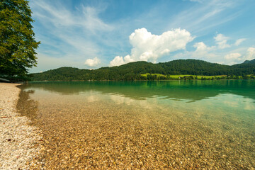 The crystal-clear waters of an alpine lake in Bavaria, Germany, reveal a pebbled bottom and reflect the surrounding green hills and blue sky with fluffy clouds, creating a serene and inviting scene.