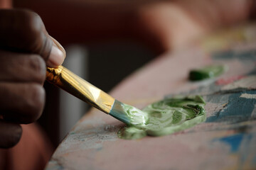 Fototapeta premium Hand holding paintbrush while dipping into green paint, adding details to canvas during artistic process