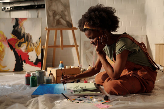 Artist sitting on floor painting with various art supplies around. Abstract and portrait paintings on easel and wall in background enhancing creative atmosphere - Powered by Adobe
