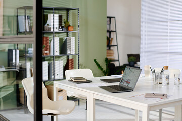 Modern office with white desk featuring open laptop, notebooks, and potted plants on shelves. Sunlight filtering through the blinds illuminating workspace