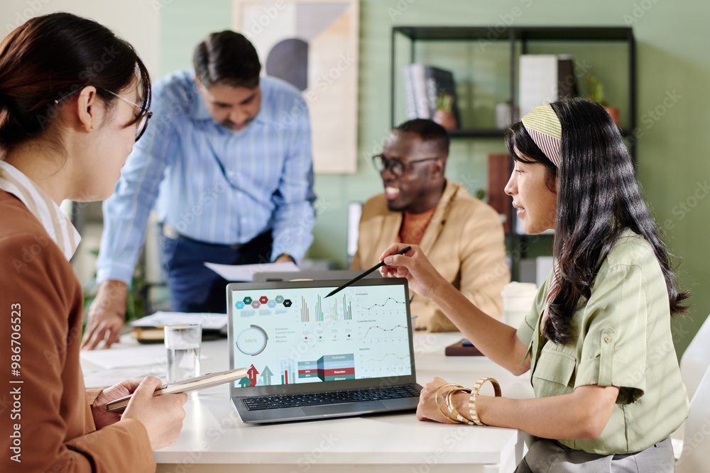 Aufkleber Multi-ethnic team discussing business analytics on laptops sitting around conference table, sharing ideas and analyzing data. Professional setting with open atmosphere and modern office design