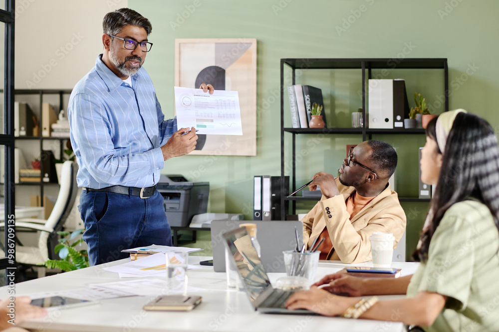 Poster middle-aged man with glasses showing charts on printed paper during business meeting while colleague