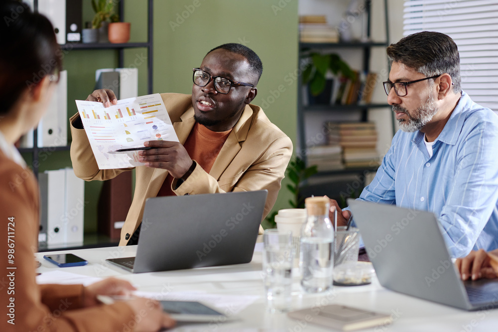 Aufkleber Business professionals sitting around table discussing performance metrics with one holding chart and another holding document showing data analysis results