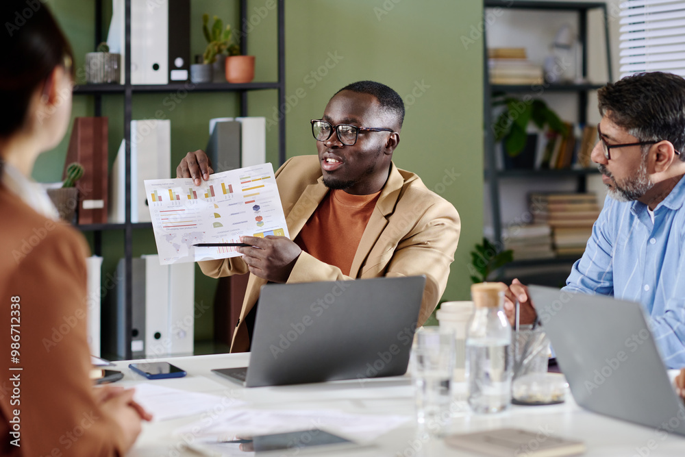 Poster Group of diverse professionals discussing and analyzing detailed business reports during meeting held in modern office with shelves and files in background