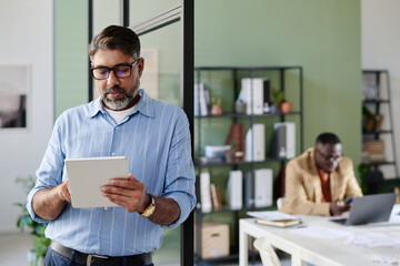 Hispanic senior man using tablet while leaning against glass wall in modern office environment...