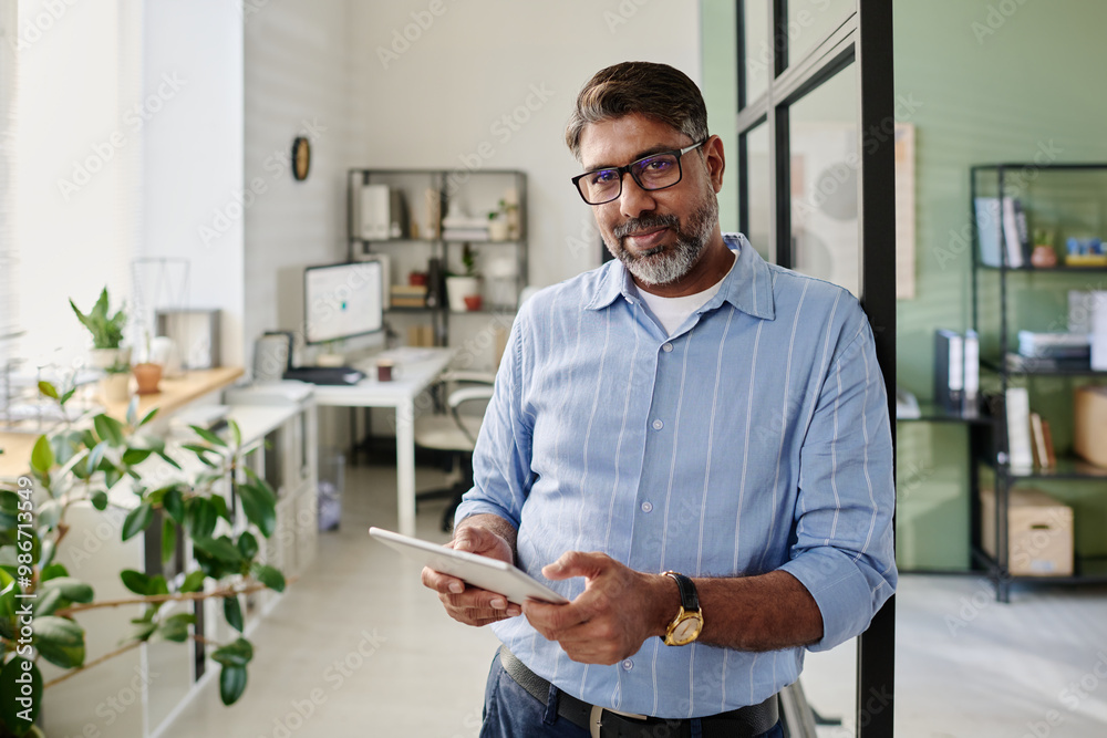 Poster portrait of bearded man in blue shirt holding tablet while standing in modern office setting with sh
