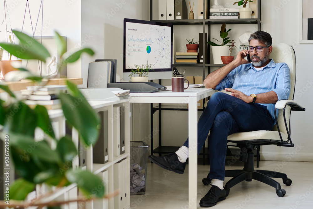 Poster Portrait of middle-aged man wearing glasses sitting at desk in office setting surrounded by computer monitors and files using smartphone for conversation