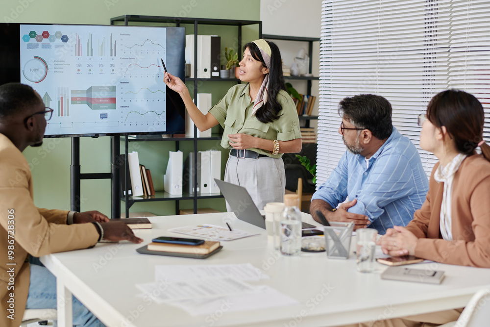 Leinwandbilder Group of colleagues discussing business data in meeting room with charts on screen and various documents on table. Presenter explaining graphs while others listening attentively