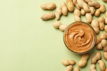 Tasty peanut butter in bowl and groundnuts on pistachio color background, flat lay. Space for text